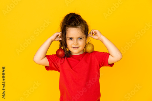 A cute little girl in a red t-shirt holding Christmas tree toys. yellow background, space for text