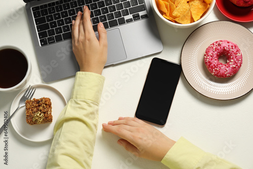 Bad eating habits. Woman using laptop surrounded by different snacks at white table, top view