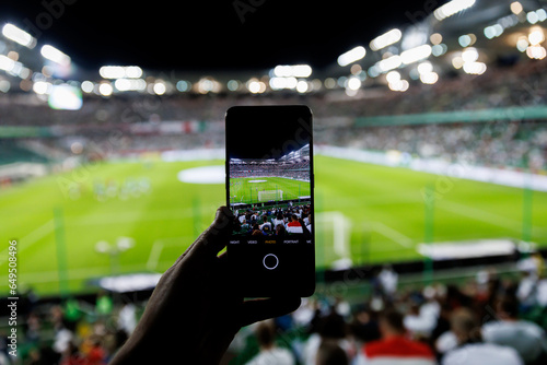 Fototapeta Naklejka Na Ścianę i Meble -  Fan hand with smartphone photographing football match. Using mobile phone camera at the stadium.