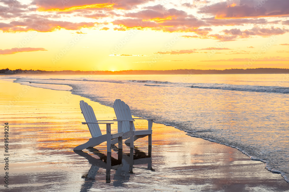 Two white chairs in the water on the ocean at dawn. Reflection of the sky on the sandy shore.