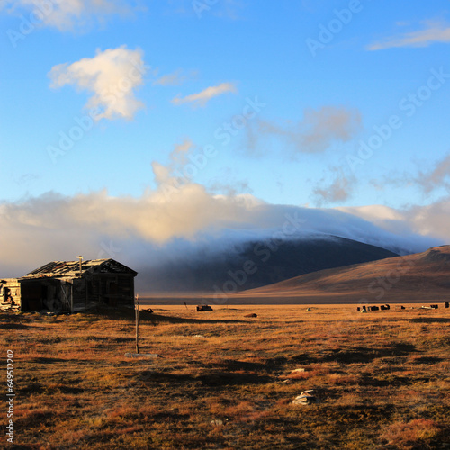 Misty mountains at sunset, a ruined old house in the Arctic tundra, autumn landscape on island, yellow grass, metal barrels on field, cloudy autumn sky, mountain foot