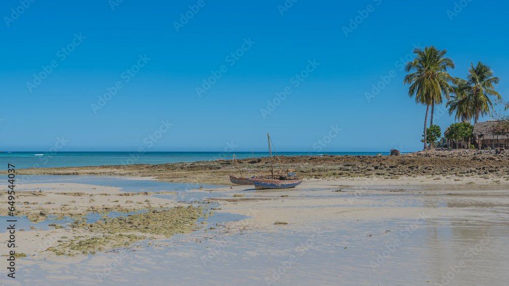 Old homemade wooden boats with masts stand on the sand at low tide. Palm trees against a clear blue sky. The ocean is far away. Reflection in puddles of water. Madagascar. Nosy Iranja