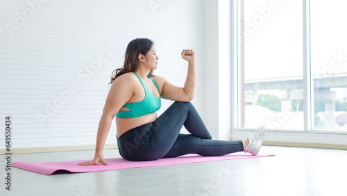 Asian young sexy chubby fat unhealthy oversized female model in sports bra legging sneakers sitting stretching arms legs on yoga pilates mat ready for training exercise practicing in home fitness gym