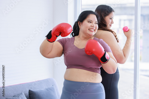 Asian young chubby fat healthy oversized overweight fit female sportswoman in casual sportswear and boxing gloves standing on yoga mat smiling punching air exercising training in home fitness gym