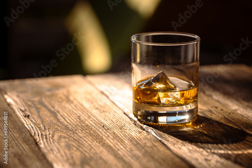 Glass with whisky with ice on a textured wooden table in harsh sunlight.