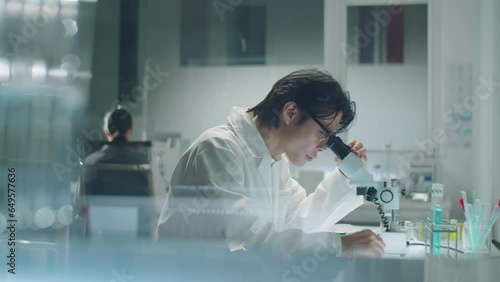 View through glass wall of Asian scientist in lab coat sitting at desk, looking through microscope and writing down notes during laboratory research