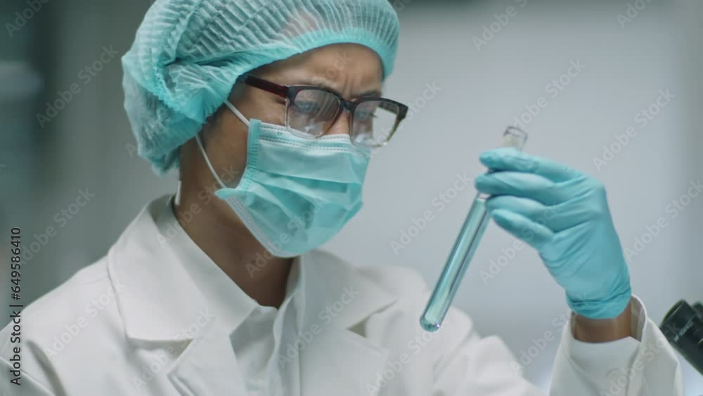 Chest up shot of Asian scientist in protective face mask, medical hat and gloves holding test tube and examining blue chemical in it while working in laboratory Chest up shot of Asian scientist in protective face mask, medical hat and gloves holding test tube and examining blue chemical in it while working in laboratory