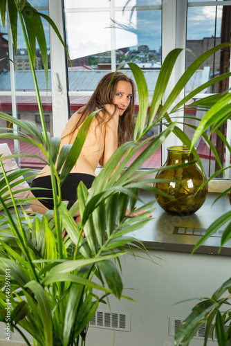 bare-breasted woman wearing black panti-slip kneels on the sill in the conservatory photo