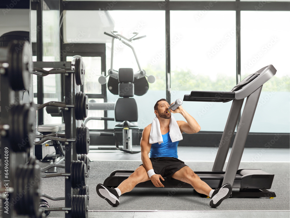 Young man drinking from a bottle and sitting on a treadmill Young man drinking from a bottle and sitting on a treadmill