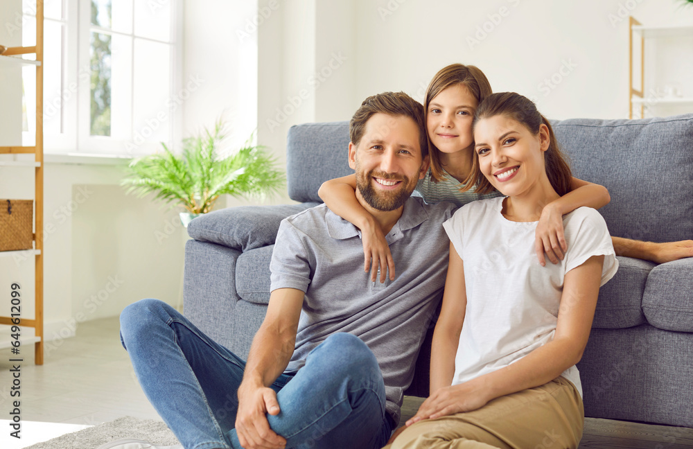 Young happy family of three sitting on the floor at home with their child girl looking at camera and smiling. Jouful daughter hugging her parents. Love, care and family leisure concept. 