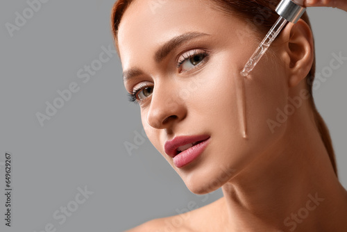 Beautiful young woman applying cosmetic serum onto her face on grey background, closeup. Space for text