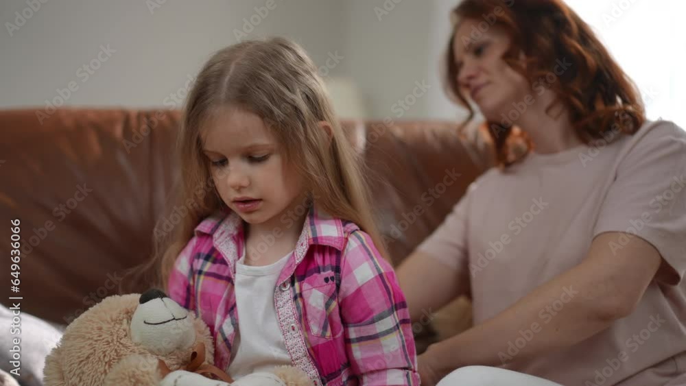 Obedient little girl playing with teddy bear as woman combing long hair. Portrait of calm relaxed Caucasian daughter with toy and loving mother taking care of child at home Obedient little girl playing with teddy bear as woman combing long hair. Portrait of calm relaxed Caucasian daughter with toy and loving mother taking care of child at home