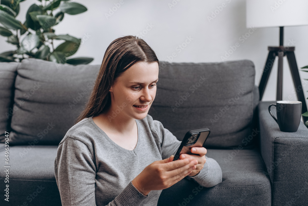 Lovely smiling young caucasian female sitting on warm floor near cozy couch at home, drinking tea or coffee, using mobile phone. Smartphone application market, podcast listening. Relaxation concept Lovely smiling young caucasian female sitting on warm floor near cozy couch at home, drinking tea or coffee, using mobile phone. Smartphone application market, podcast listening. Relaxation concept