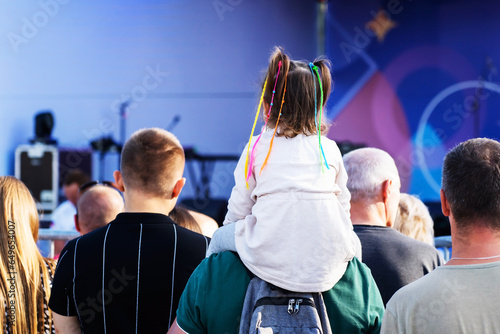 A little girl with colorful pigtails sits on her father's shoulders during a mass outdoor concert. A crowd of people watches a show at a concert venue. Unrecognizable person. Foreground