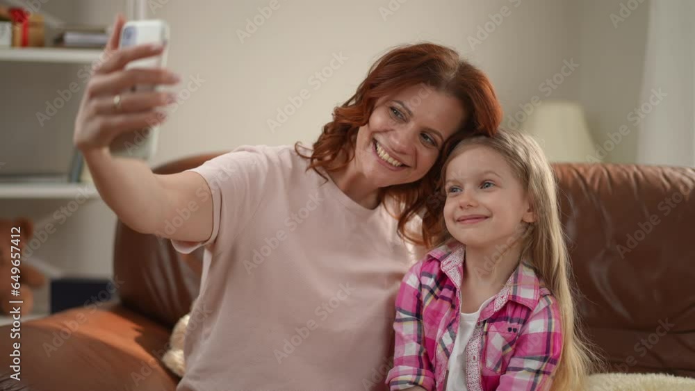 Happy Caucasian mother and daughter taking selfie with smartphone app sitting at home in living room. Portrait of positive woman and girl smiling photographing indoors Happy Caucasian mother and daughter taking selfie with smartphone app sitting at home in living room. Portrait of positive woman and girl smiling photographing indoors