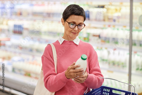 Waist up portrait of mature woman holding bottle of milk in supermarket and looking at expiration date, copy space