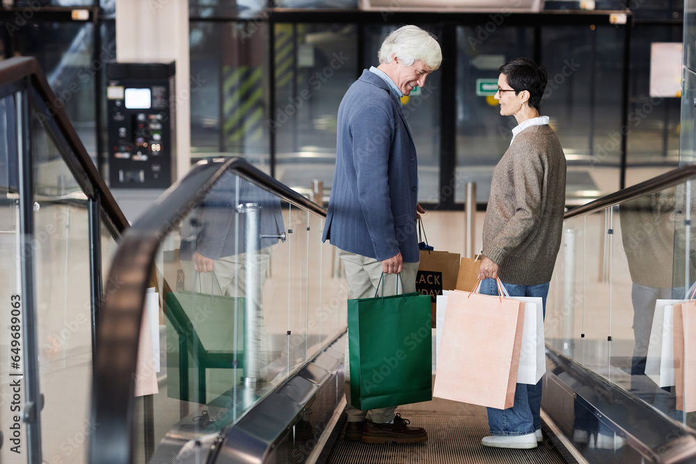 Side view of senior couple holding shopping bags standing on escalator in shopping mall, copy space