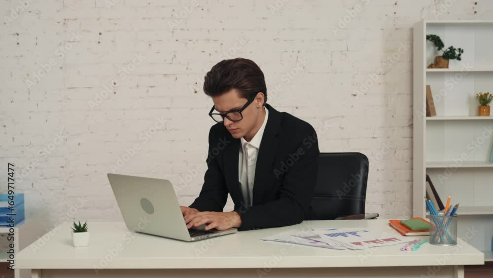 Medium video of a young man sitting in the office at the table, working on a laptop. He takes his glasses off, rubs his eyes, exhausted. Medium video of a young man sitting in the office at the table, working on a laptop. He takes his glasses off, rubs his eyes, exhausted.