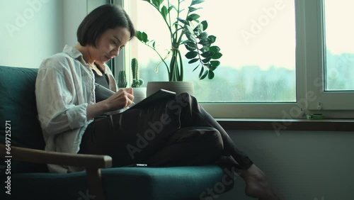 Young woman artist drawing on digital tablet. Creative girl working on tablet with digital pencil. Artist using computer to create art sitting near window 