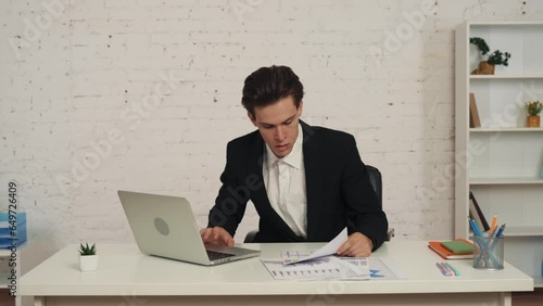 Medium video of a young man sitting in the office, working on a laptop and cheching the graphs and charts on his table. He loooks confused and stressed. photo