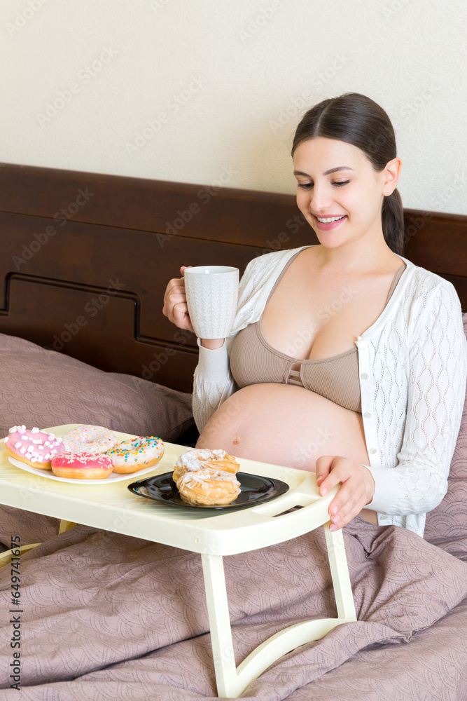 Pregnant woman is having breakfast in bed at home. Tray with donuts and cakes. Eating pastry during pregnancy concept
