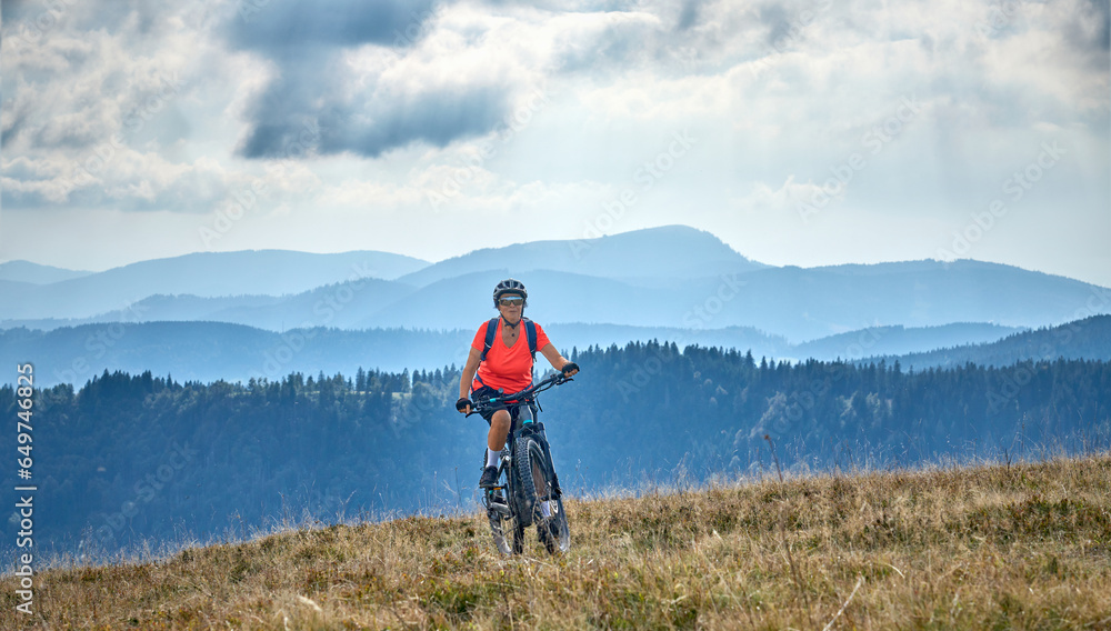 Fototapeta premium nice senior woman on her electric mountain bike cycling on Feldberg summit with stunning view over the Black Forest mountains and valleys, Baden-Wuerttemberg Germany