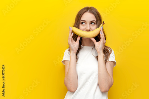 young girl shows smile and holds banana on yellow isolated background, woman with fruit looks at copy space