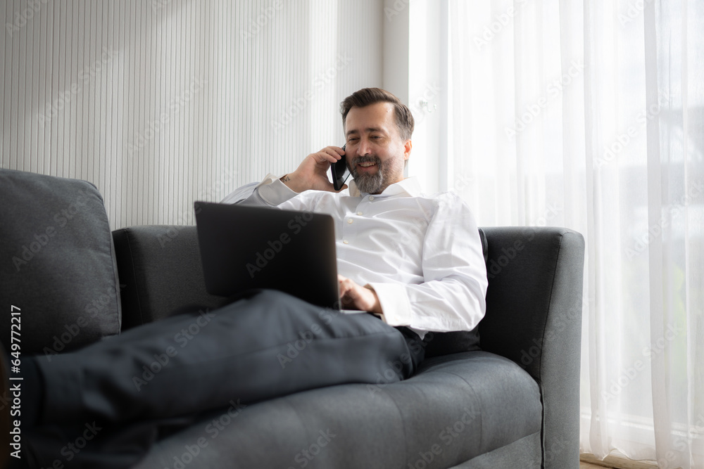 Businessman using laptop and talking on the phone while sitting on sofa at home
