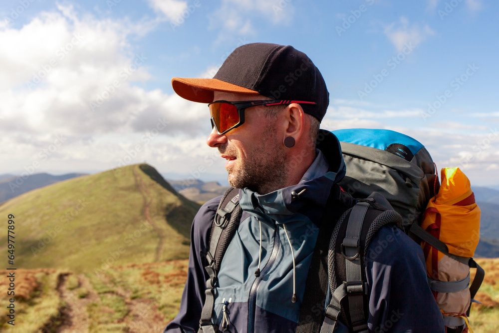 man in jacket and sunglasses stands on the mountain and looks at the sun, tourist guy in mountain clothes