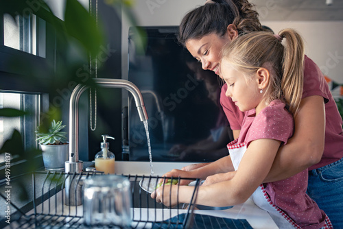 A cute little girl helps her mother wash dishes. Child doing house chores. Mother teaches a child to wash dishes.