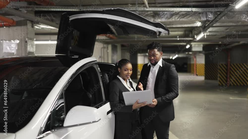 African working mates in formal outfits looking at laptop screen in underground garage with e-car. Commercial partners inspecting financial report via modern technologies in bottom-level parking. African working mates in formal outfits looking at laptop screen in underground garage with e-car. Commercial partners inspecting financial report via modern technologies in bottom-level parking.