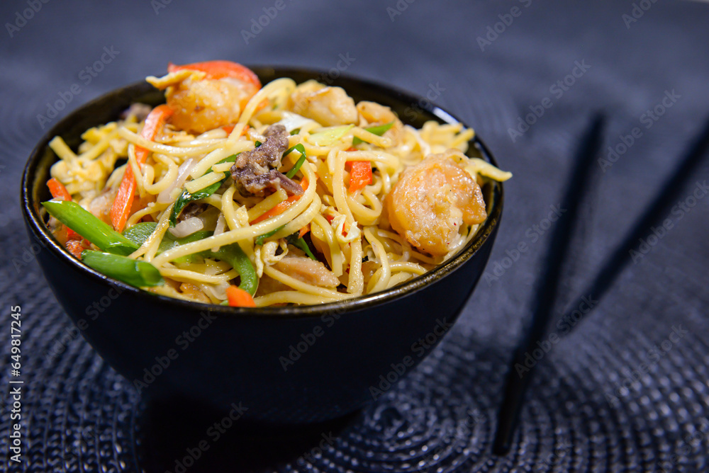 mixed fried noodles with with chicken, carrot, spring onion, bell pepper, egg and chopsticks served in bowl side view of chinese food mixed fried noodles with with chicken, carrot, spring onion, bell pepper, egg and chopsticks served in bowl side view of chinese food