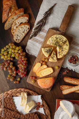 cheese on a wooden board surrounded by bread, grapes, crackers and spices on a wooden table, top view