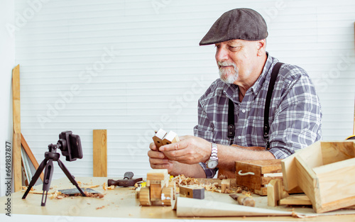 Senior old Caucasian man wearing check shirt, apron, making DIY wooden furniture, using mobile phone, streaming live video clip, selling product in social media at home. Retirement, Hobby Concept. photo