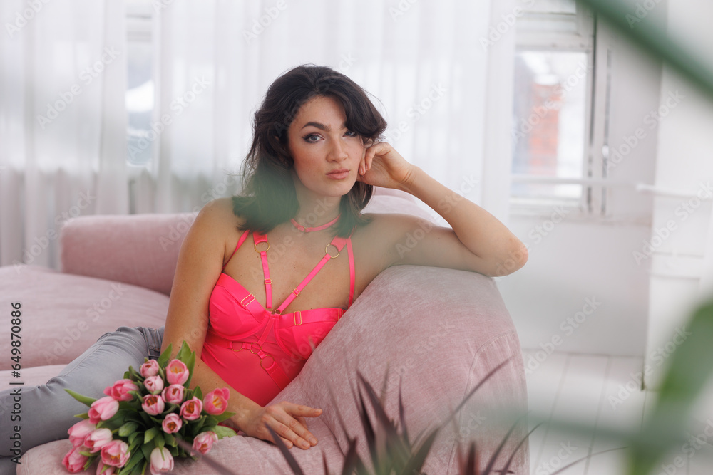 Beautiful brunette woman sitting on pink velvet sofa with a bucket of flowers tulips. Girl in pink bodysuit near window. Portrait of young woman smile. Living room. Beautiful brunette woman sitting on pink velvet sofa with a bucket of flowers tulips. Girl in pink bodysuit near window. Portrait of young woman smile. Living room.