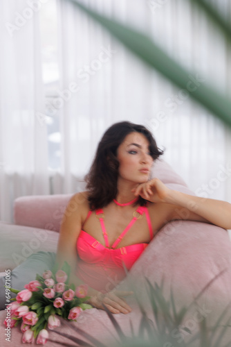 Beautiful brunette woman sitting on pink velvet sofa with a bucket of flowers tulips. Girl in pink bodysuit near window. Portrait of young woman smile. Living room.