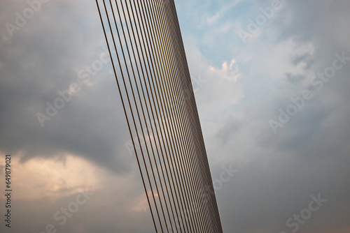 A suspension bridge cable, composed of smaller cables, extends diagonally against a cloudy sky.