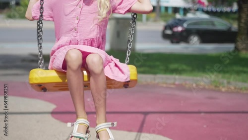 Blonde little girl rides swings with chains in city park smiling child in casual dress plays attraction on urban playground schoolgirl swings in sunny garden photo