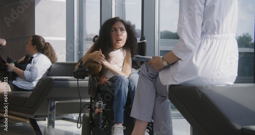 Woman with SMA in electric wheelchair talks to doctor in hospital or clinic lobby. Female physician consults patient with physical disability. People sit on sofa in modern medical center waiting area.