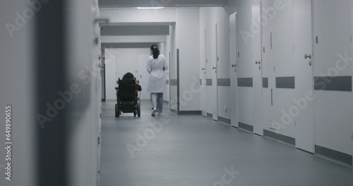 Female doctor walks down the clinic hallway, consults woman with physical disability. Physician talks about medical procedures with patient in electric wheelchair. Corridor of modern medical facility.