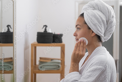 Woman cleaning facial skin with cotton disk near mirror in bathroom beautiful lady with towel turban taking care of skin with dermatological product at dressing table at home photo