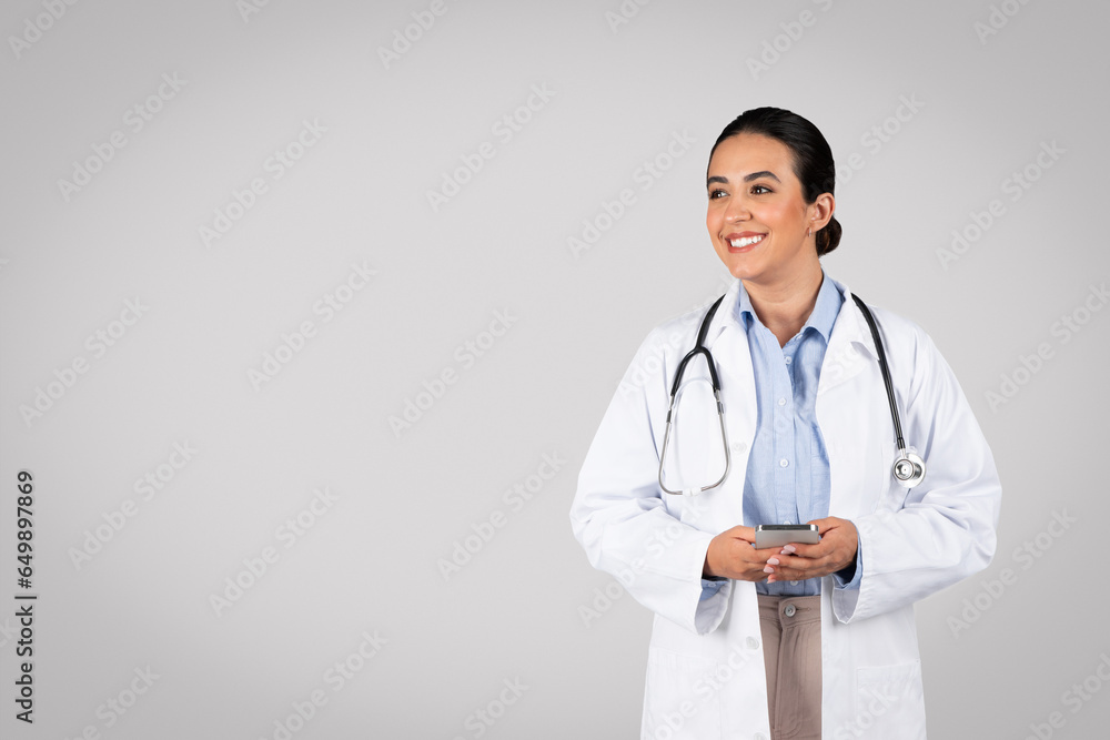 Happy brazilian woman doctor in white coat using cellphone and looking aside at free space, giving online consultation Happy brazilian woman doctor in white coat using cellphone and looking aside at free space, giving online consultation