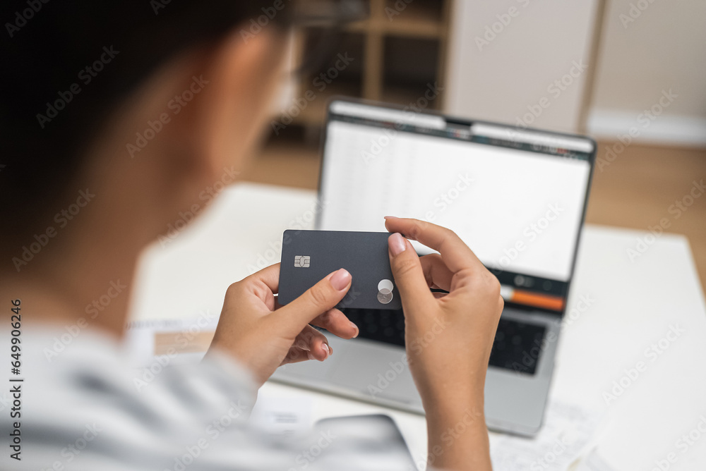 Lady with dark hair holds black credit card in hands against switched laptop woman paying for utilities via bank online system on modern device from home