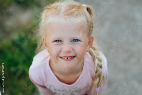 Close up portrait of blonde caucasian little girl looks up at camera smiles. Childhood, outdoor leisure, vacations. Preschooler girl with braid enjoying nature at country house.