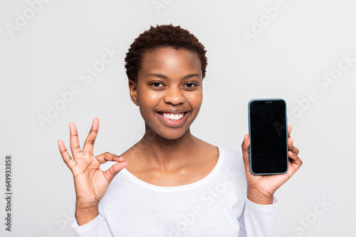 Banner advertisement concept delighted dark skinned african woman with short haircut smiing showing okay sign holding cellphone smartphone in hands with bleck empty screen copy space photo