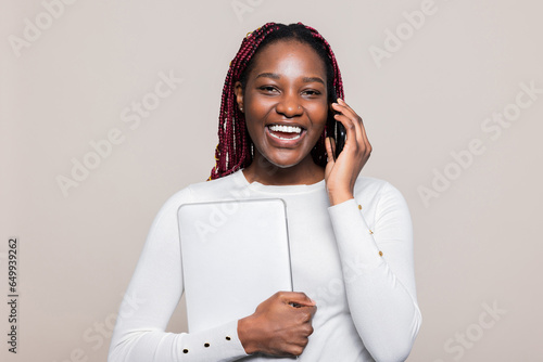 Joyful African American woman with vibrant braids is speaking on the phone with classmates friends while smiling at the camera preparing to go to University High school holding digital laptop in hand