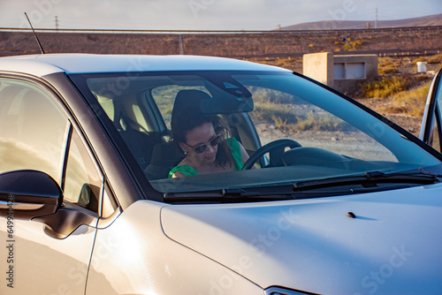 White woman in sunglasses getting into the white car preparing her seat to start driving in a dry brown mountains landscape at sunset.