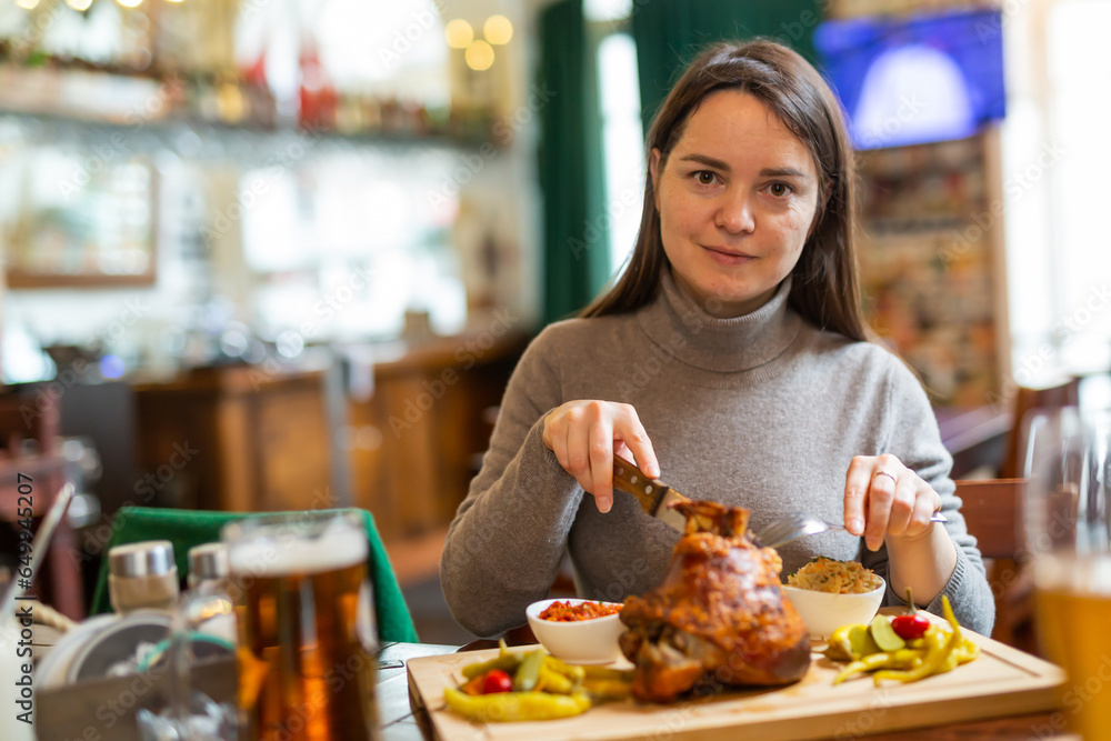 Female traveler tasting fried pork ham knuckle traditionally served with stewed cabbage, pickled vegetables, various sauces and beer in cafe in Vienna. Popular snacks