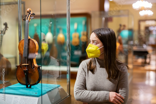 Interested young woman wearing protective face mask viewing collections of ancient musical instruments in anthropological museum. Forced precautions in pandemic photo