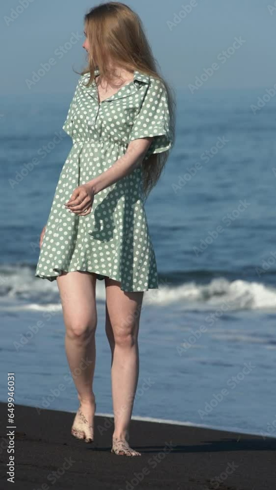Attractive blonde woman 21-year-old walking along beach of black sand on Pacific Coast. Cheerful female dressed in summer green polka dot dress, barefoot. Vertical slow motion, handheld. Panning right Attractive blonde woman 21-year-old walking along beach of black sand on Pacific Coast. Cheerful female dressed in summer green polka dot dress, barefoot. Vertical slow motion, handheld. Panning right